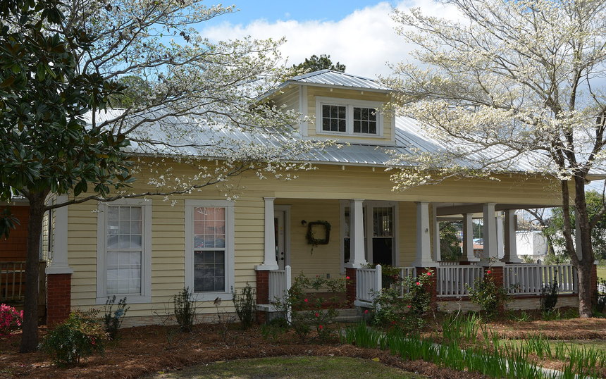A yellow bungalow with a deep front porch behind flowering trees